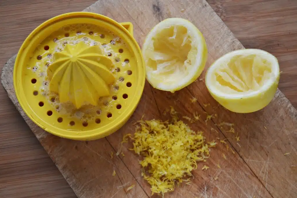A yellow citrus juicer, two squeezed lemon halves, and a pile of lemon zest are on a wooden cutting board.