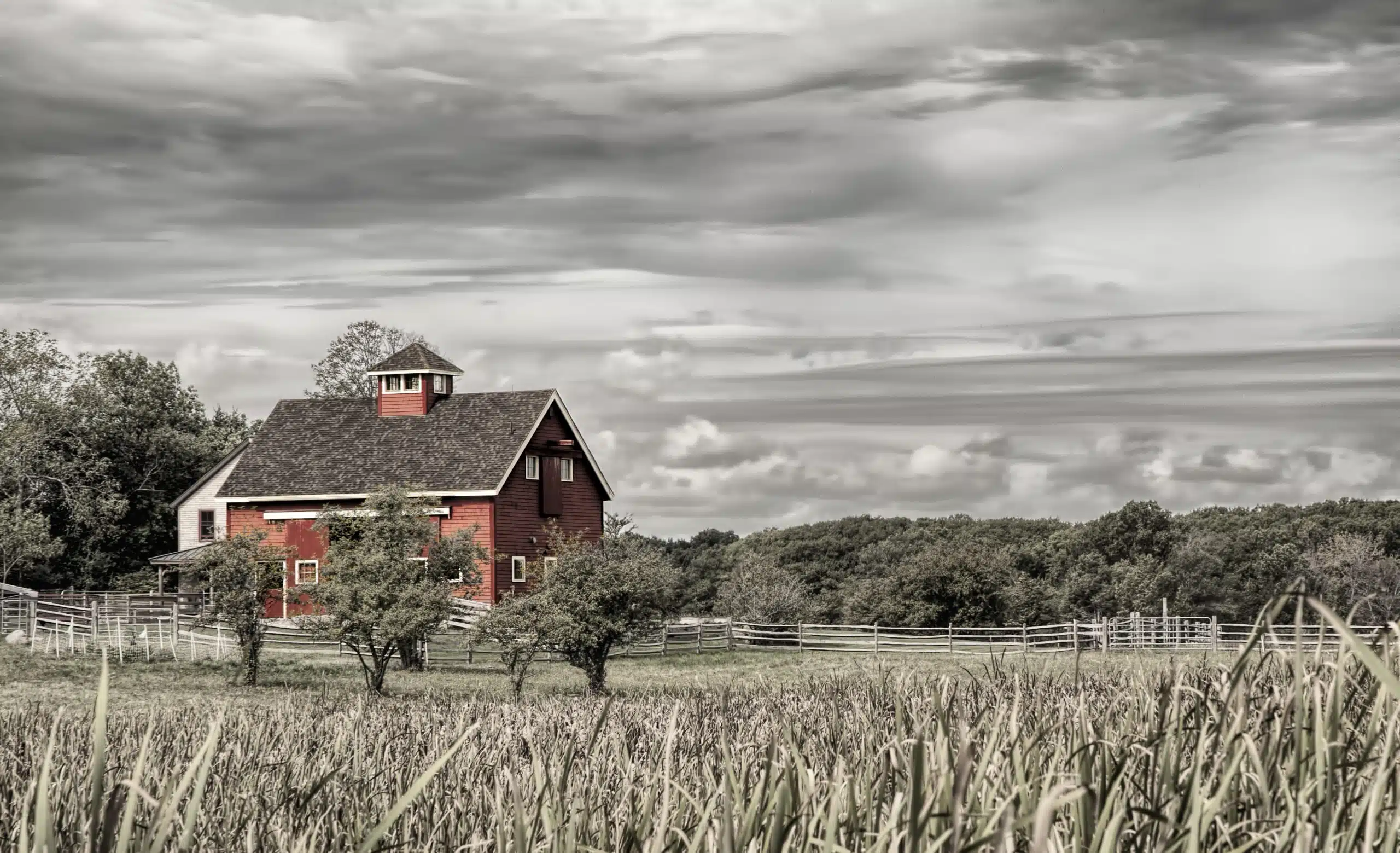 A red barn with a dark roof stands amidst green trees and fields under a cloudy sky, bordered by a wooden fence.