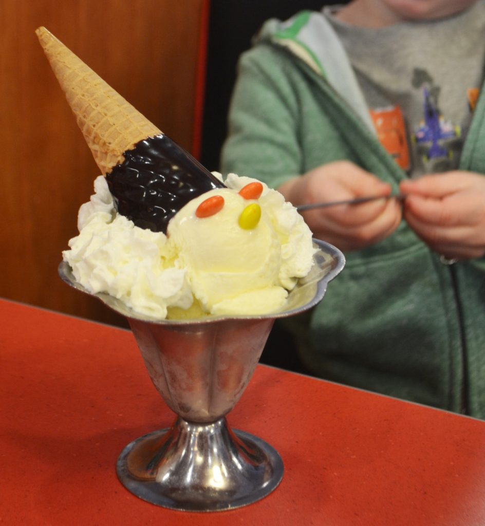 Sundae shaped like a clown's head with whipped cream, candy eyes, and a cone hat; child in background.