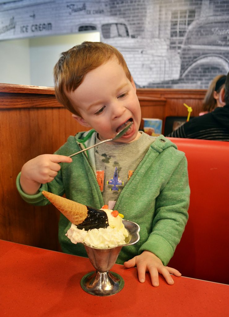 Young boy in a green hoodie eating ice cream topped with a cone and chocolate sauce.