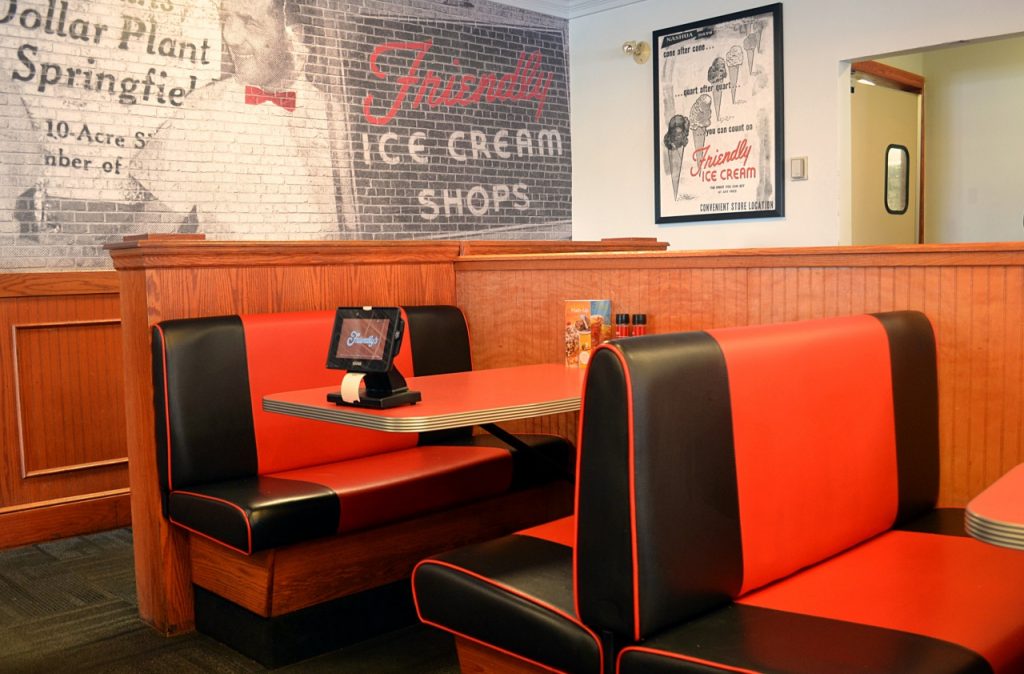 A vintage-style ice cream shop interior with red and black booths, wood paneling, and a brick wall mural featuring ice cream ads. A table is set with a menu, condiments, and an electronic device.