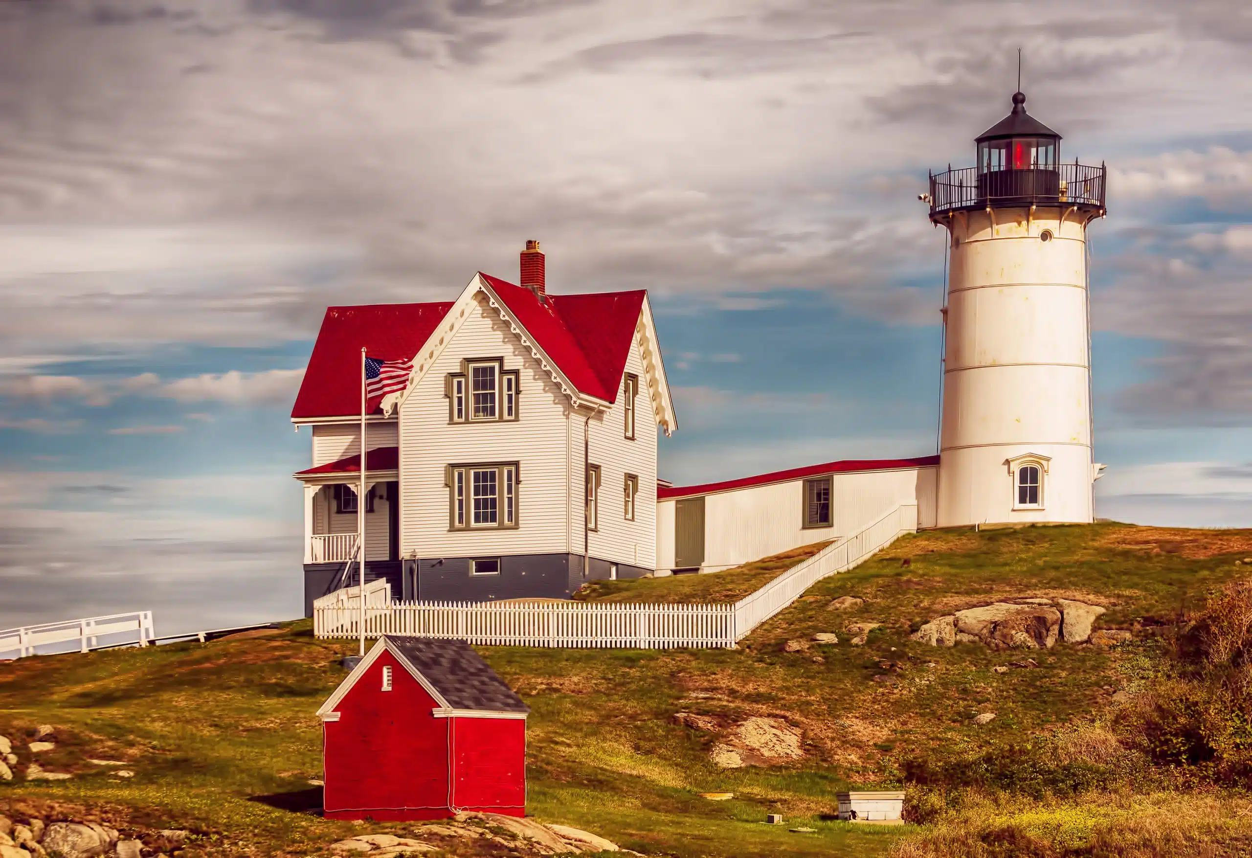 A lighthouse with an attached white house featuring a red roof stands on a grassy hill next to a smaller red building. A white fence surrounds part of the area. The sky is cloudy.