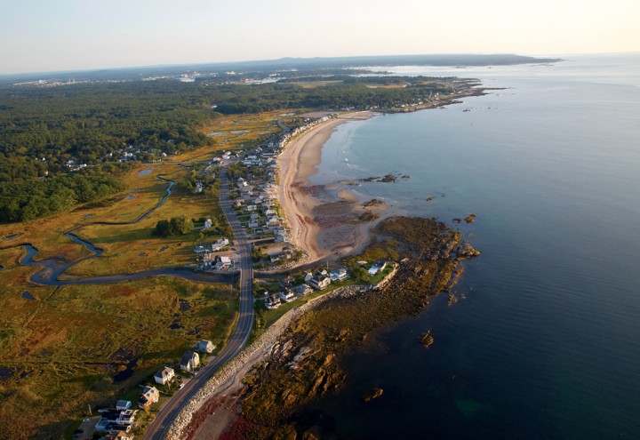 Looking north at sunrise, an aerial view catches Rye’s Ocean Boulevard tracing the New Hampshire coastline behind Concord Point and the beach at Wallis Sands State Park. To the west of the roadway are protected marshlands, drained by Parsons Creek.