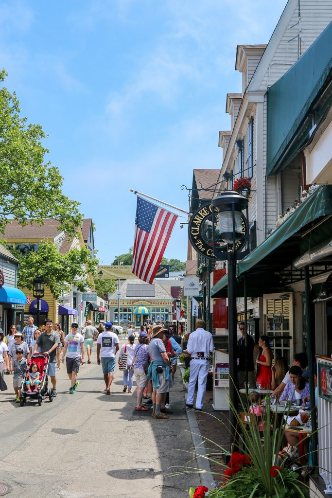 A bustling street scene with people walking, an American flag hanging, and shops lined on both sides of the road.