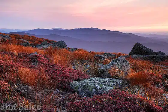 A rocky landscape with autumnal red and orange vegetation is seen under a colorful sky at sunset, with mountains in the background.