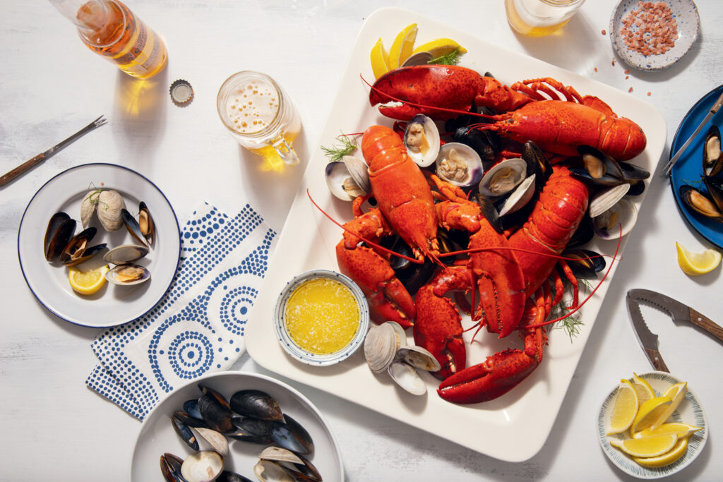 A platter of cooked lobster, clams, and mussels is served with lemon wedges, a bowl of melted butter, and drinks on a white table.
