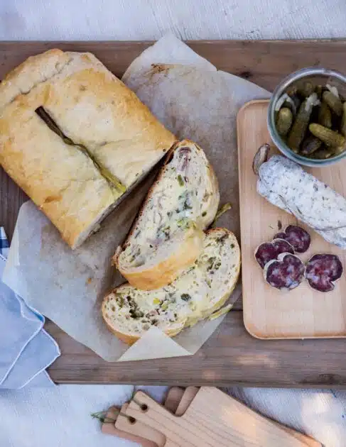 Sliced savory loaf, cured sausage, pickles, and napkin on a wooden table with parchment paper.