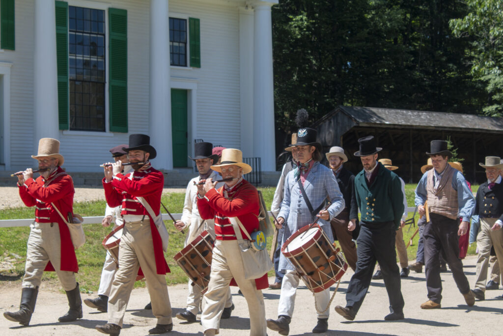 Men in historic costumes march and play drums and flutes near a white building with green shutters.