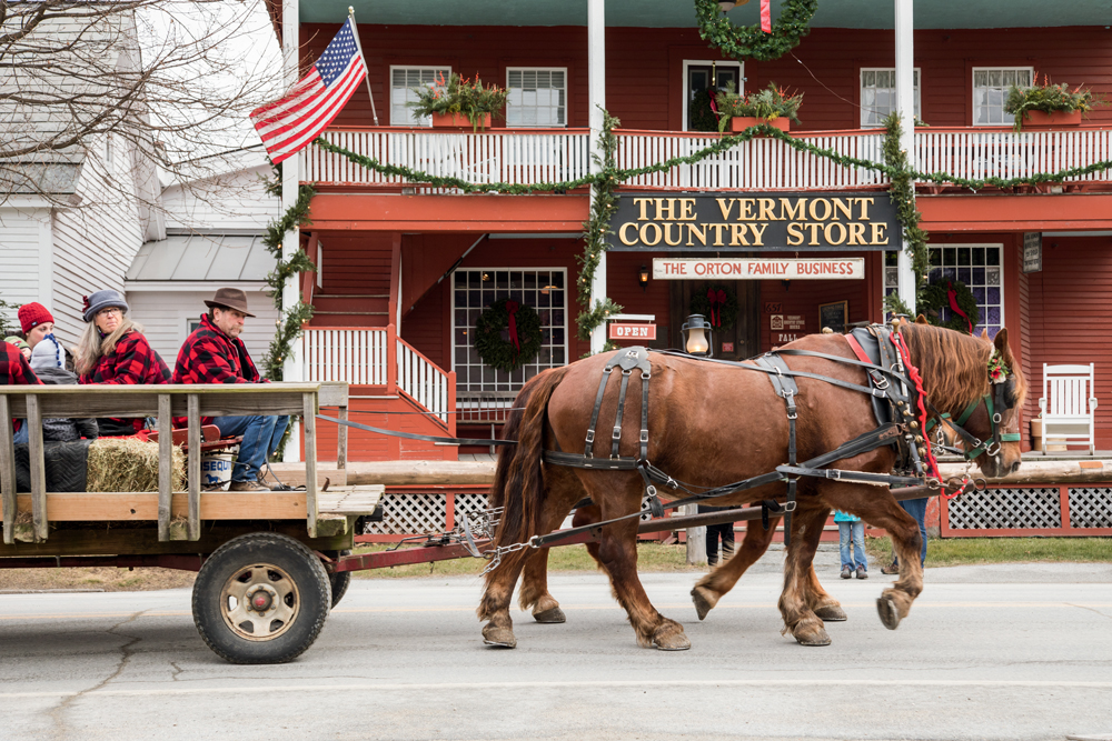 Two large horses pull a wagon with people sitting on hay bales in front of The Vermont Country Store, a red building decorated with wreaths and an American flag.