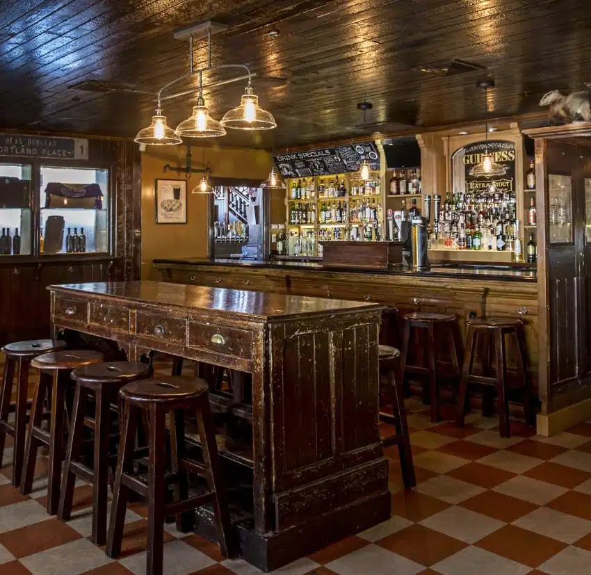 Cozy pub interior with wooden tables, bar stools, and a well-stocked bar on a checkered tile floor.