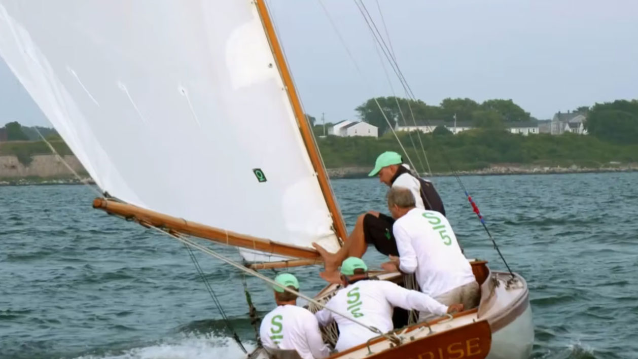 Four people wearing matching shirts and green caps are sailing a wooden sailboat on a body of water, with land and houses visible in the background.