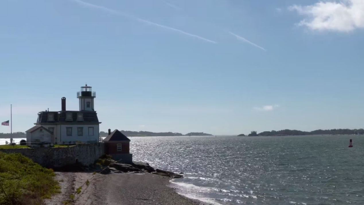 A lighthouse and adjacent buildings stand on a rocky shoreline beside a body of water under a clear sky.