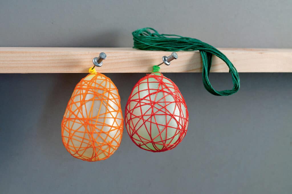 Two balloons wrapped in colored string hang from a wooden shelf, with green string coiled on top.