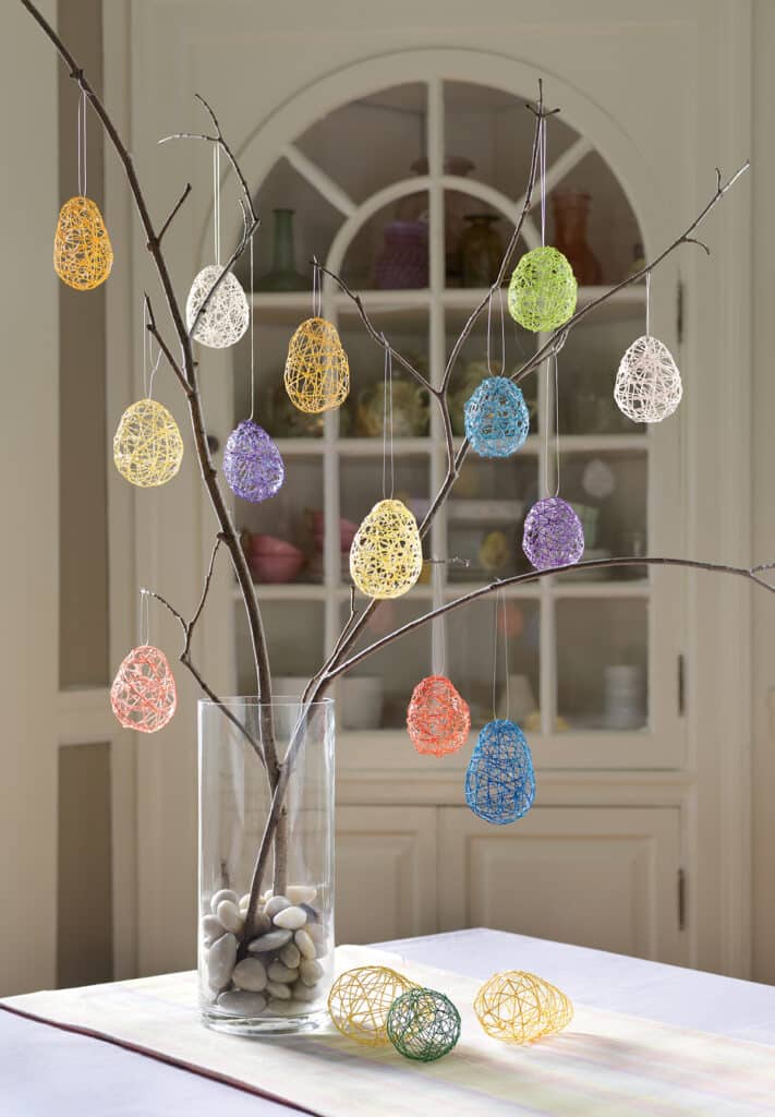 String Easter Eggs. Bare branches in a glass vase decorated with colorful string Easter egg ornaments, some eggs on the table.