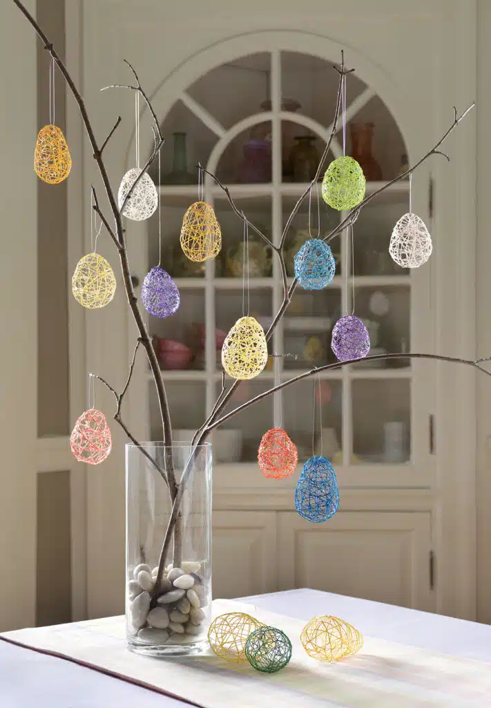 String Easter Eggs. Bare branches in a glass vase decorated with colorful string Easter egg ornaments, some eggs on the table.