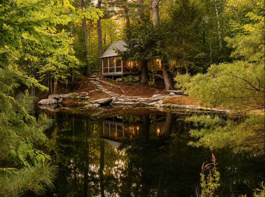 A cozy cabin with lit windows sits by a calm forest pond, surrounded by lush green trees and stone steps.