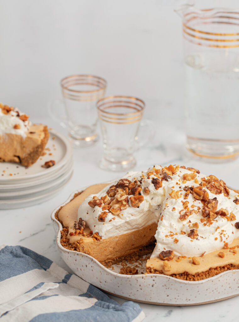 A pumpkin chiffon pie topped with whipped cream and chopped nuts sits on a table; a slice is cut and served on a plate nearby, with glasses and a pitcher of water in the background.