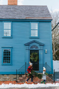 A woman and a child carrying evergreen branches walk toward the entrance of a blue house.