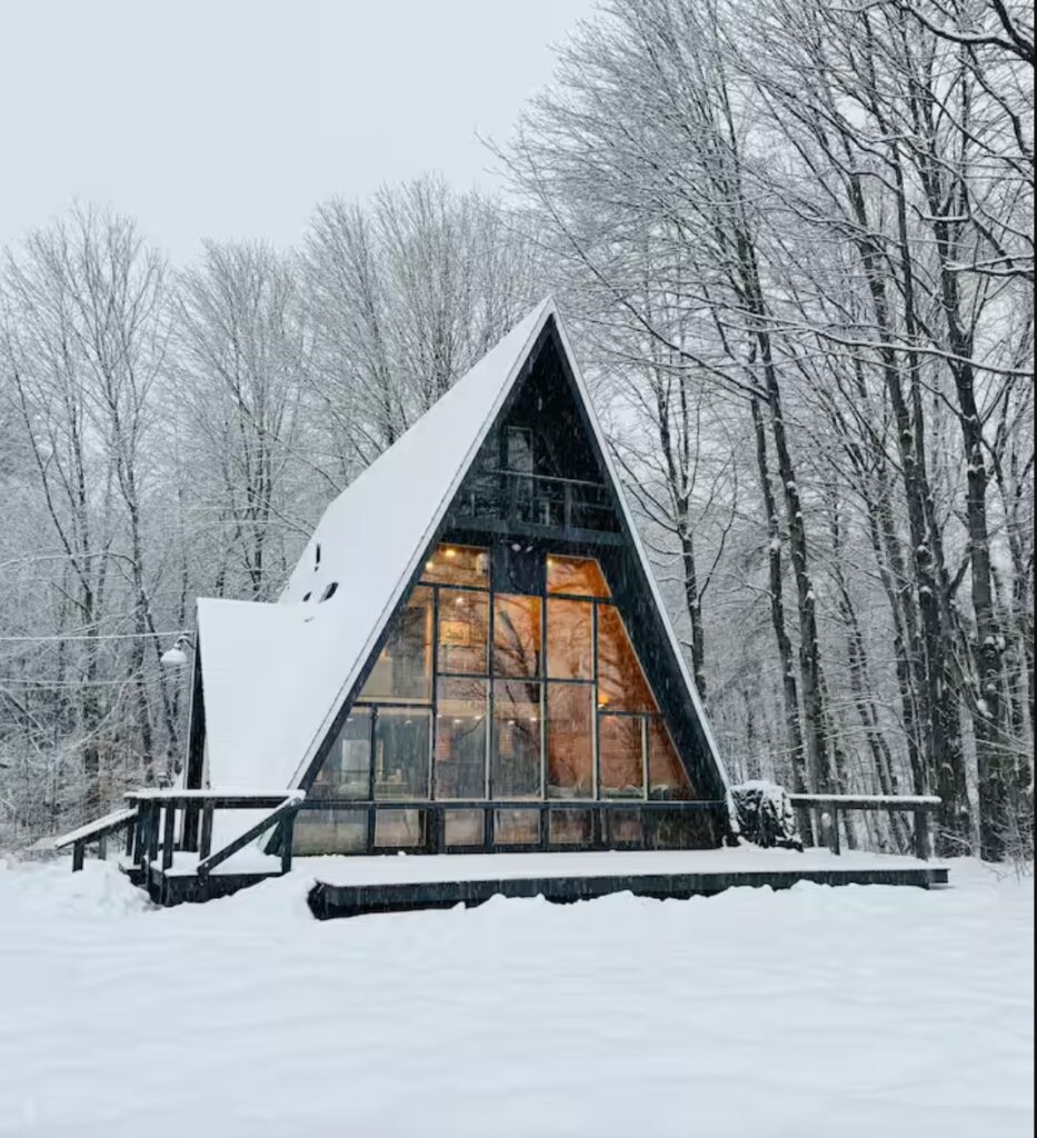 A-frame cabin with glowing windows in a snowy forest, surrounded by bare trees in winter.