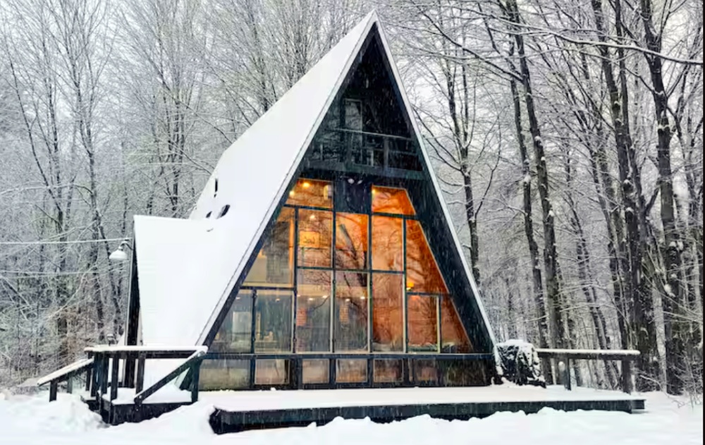 A-frame cabin with large glass windows, glowing lights inside, and snow-covered trees all around.