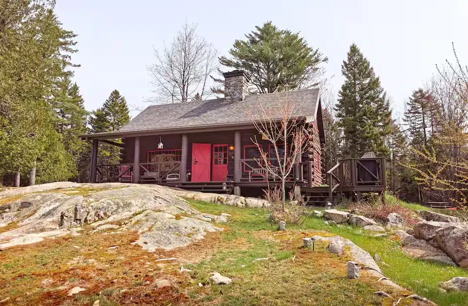 A rustic cabin with a red door sits on rocky ground, surrounded by trees and greenery.