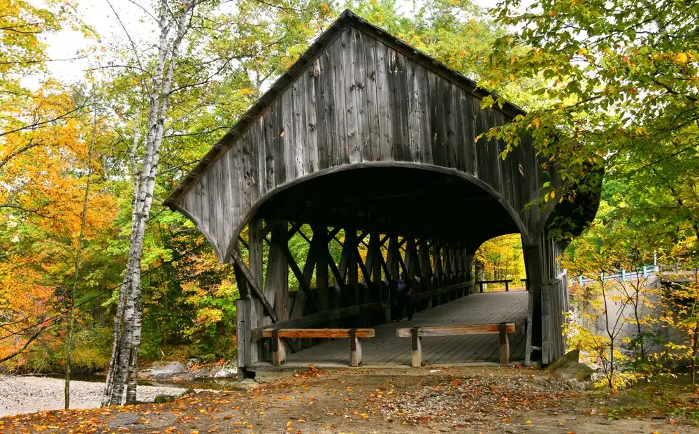 The Best Covered Bridge in Every New England State. Wooden covered bridge surrounded by colorful autumn trees, with leaves scattered on the ground.