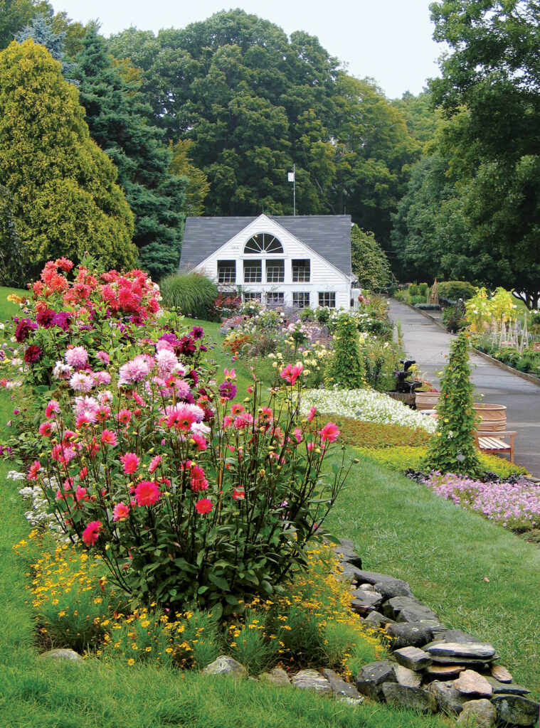A white house with large windows sits behind vibrant flower gardens and a stone path on a lush green lawn.
