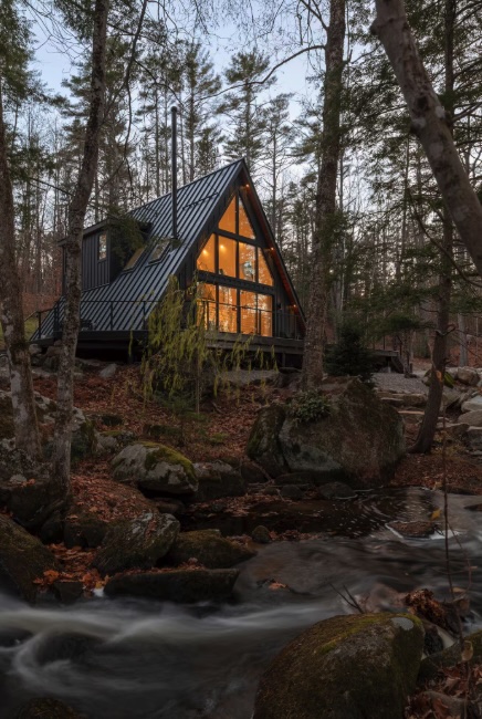 A cozy A-frame cabin with glowing windows sits among trees beside a flowing stream in a forest.