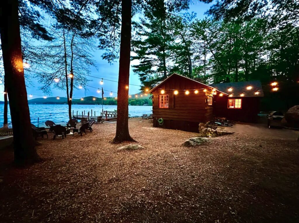 Cozy lakeside cabin at dusk with string lights, firepit, and Adirondack chairs surrounded by tall trees.