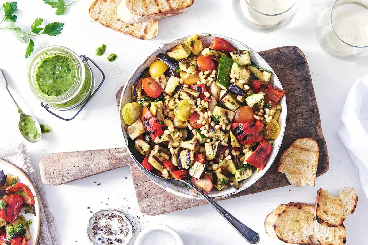 A bowl of grilled vegetable salad, surrounded by toast and a jar of pesto on a white table.
