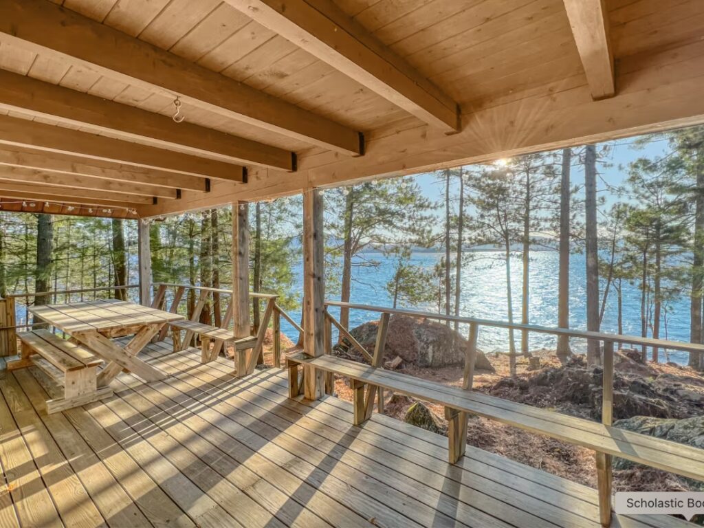Wooden cabin porch with benches and table, overlooking a lake and trees in bright sunlight.