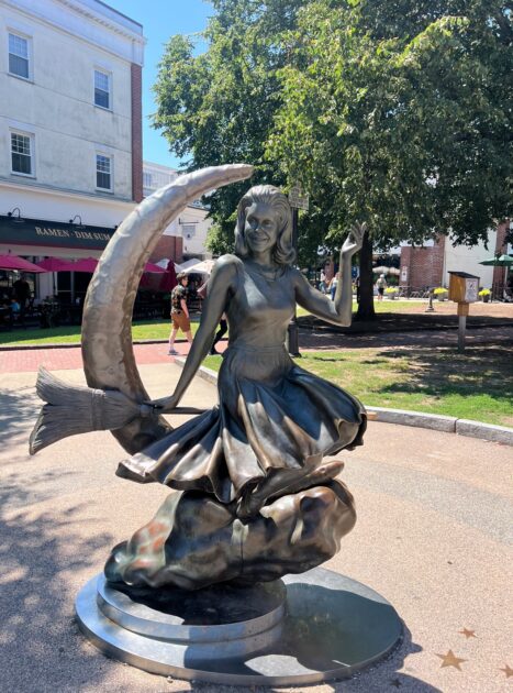 A bronze statue of a woman in a dress holding a crescent moon stands outdoors on a circular base, with trees and buildings in the background.