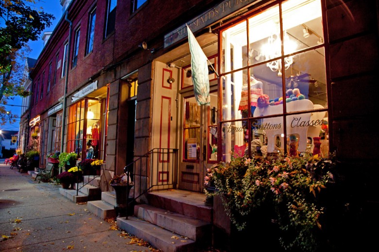 A warmly lit yarn and craft shop with a decorated window and plants outside is open on a city street in the evening, one of the best antique towns in New England.
