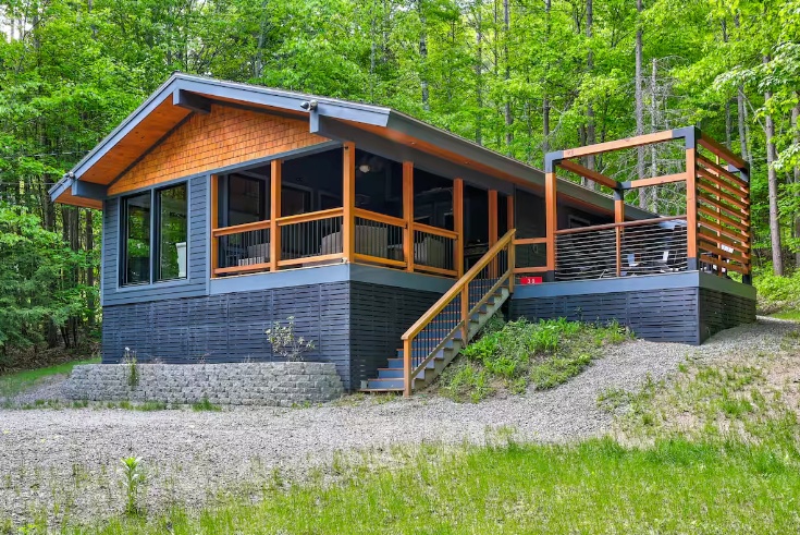 A modern cabin with wooden accents and large windows, set in a green, wooded area with a gravel path and a small porch with railings.