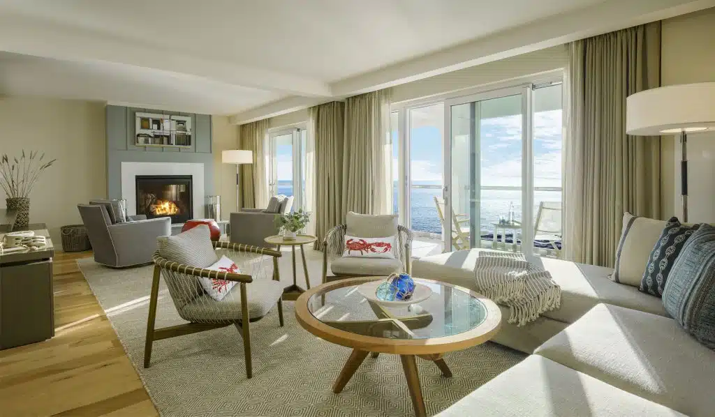 Luxury suite: Bright living room with modern furniture, fireplace, and large windows overlooking the ocean and balcony.