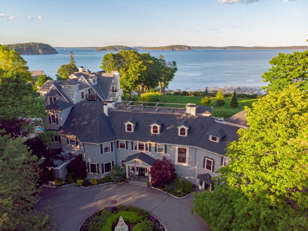 Large gray house with dormer windows, surrounded by trees, overlooking a calm bay with islands in the distance.