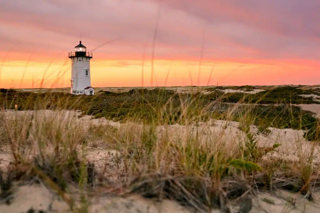 Best Massachusetts Beaches. A lighthouse stands on sandy dunes with grass in the foreground under a colorful sunset sky.
