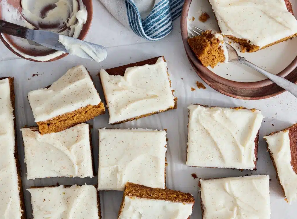 Pumpkin Sheet Cake Bars with Cream Cheese Frosting | Several rectangular pieces of cake with cream cheese frosting are arranged on a white surface, with a plate, fork, and a bowl of leftover frosting visible.