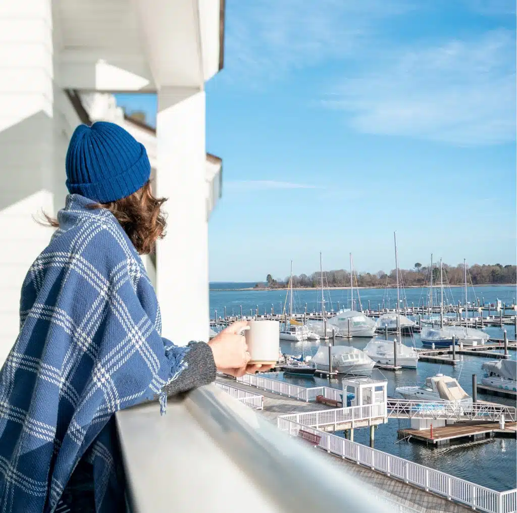 Person in blue hat and blanket holds mug, looking at marina with boats on a sunny day.