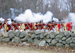 Historical reenactors in red uniforms fire muskets behind a stone wall in an outdoor setting.