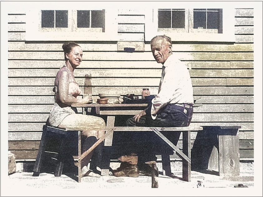 Two people sit at an outdoor wooden table eating a meal in front of a wooden building with three small windows.