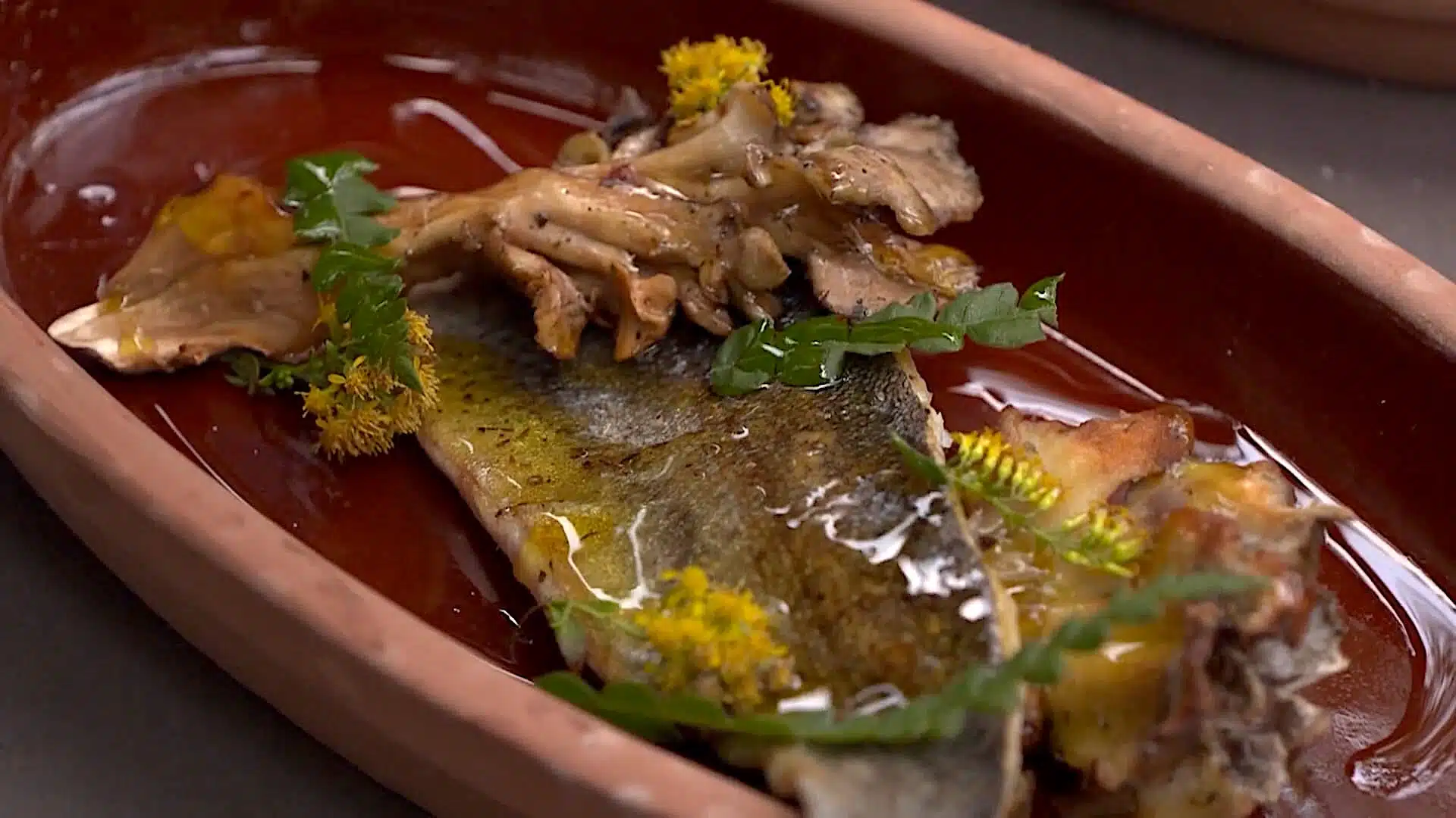 A cooked fish fillet served in a brown ceramic dish, garnished with mushrooms, leafy greens, yellow flowers, and a drizzle of oil.