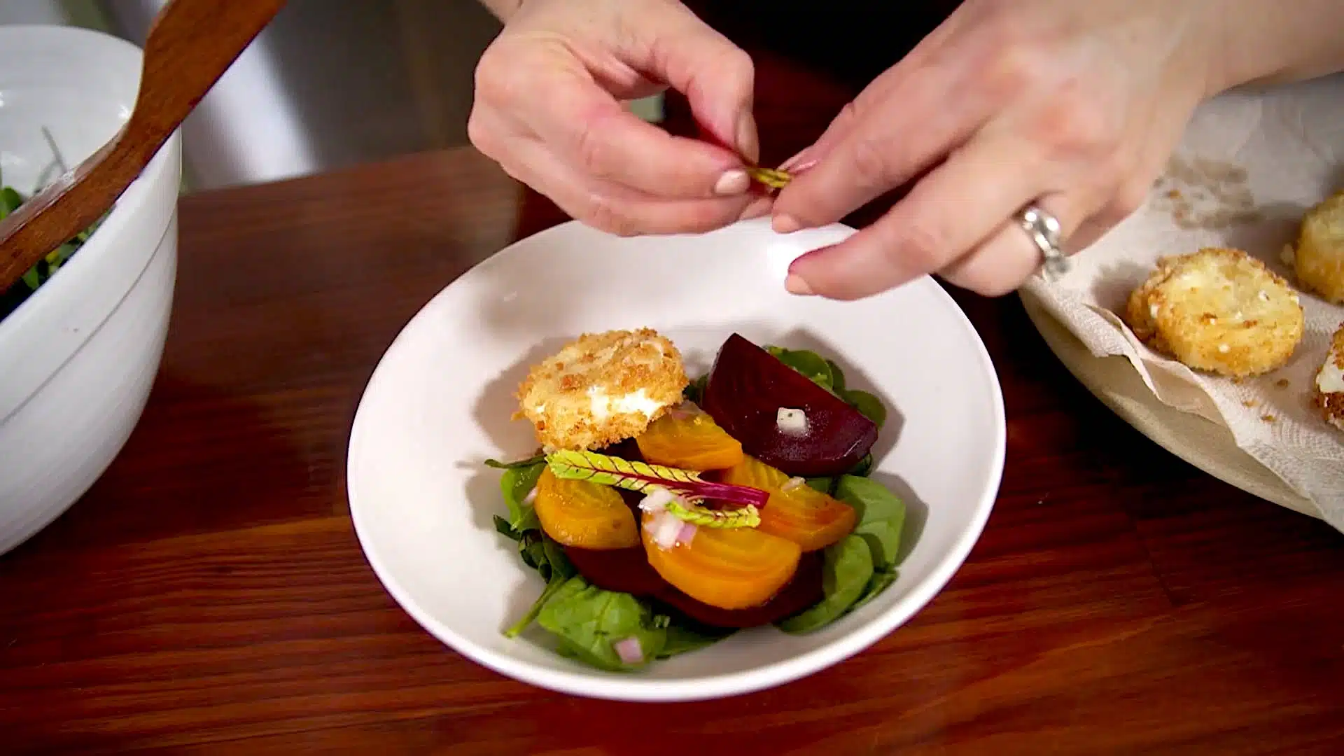 A person garnishes a bowl of beet salad with herbs; the salad includes yellow and red beets, greens, and a breaded cheese round on a wooden table.