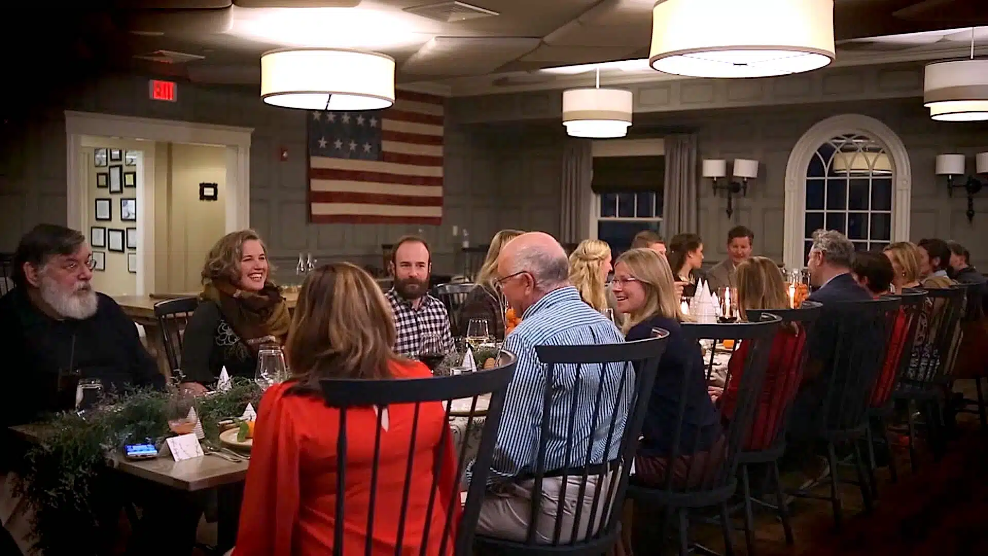 A group of people are sitting around a long dining table in a well-lit room with a U.S. flag on the wall.