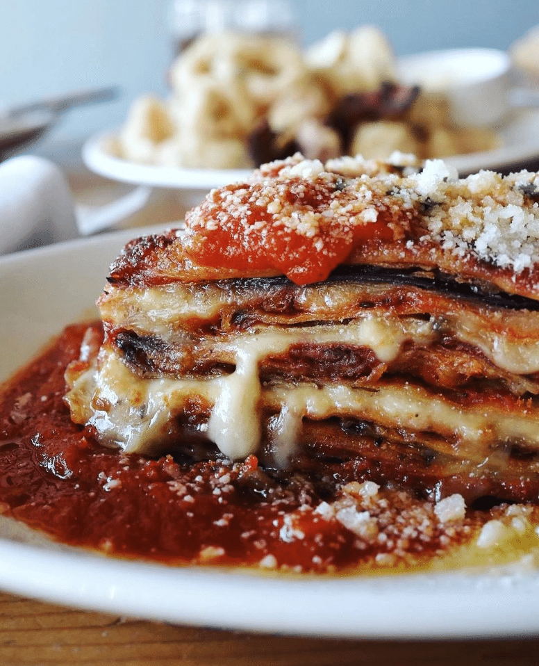 Close-up of a slice of layered chicken parmesan with layers of cheese and tomato sauce on a white plate, topped with grated parmesan cheese.