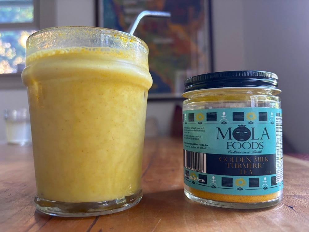 A glass of golden milk with a metal straw next to a jar of Mola Foods Golden Milk Turmeric Tea on a wooden table.