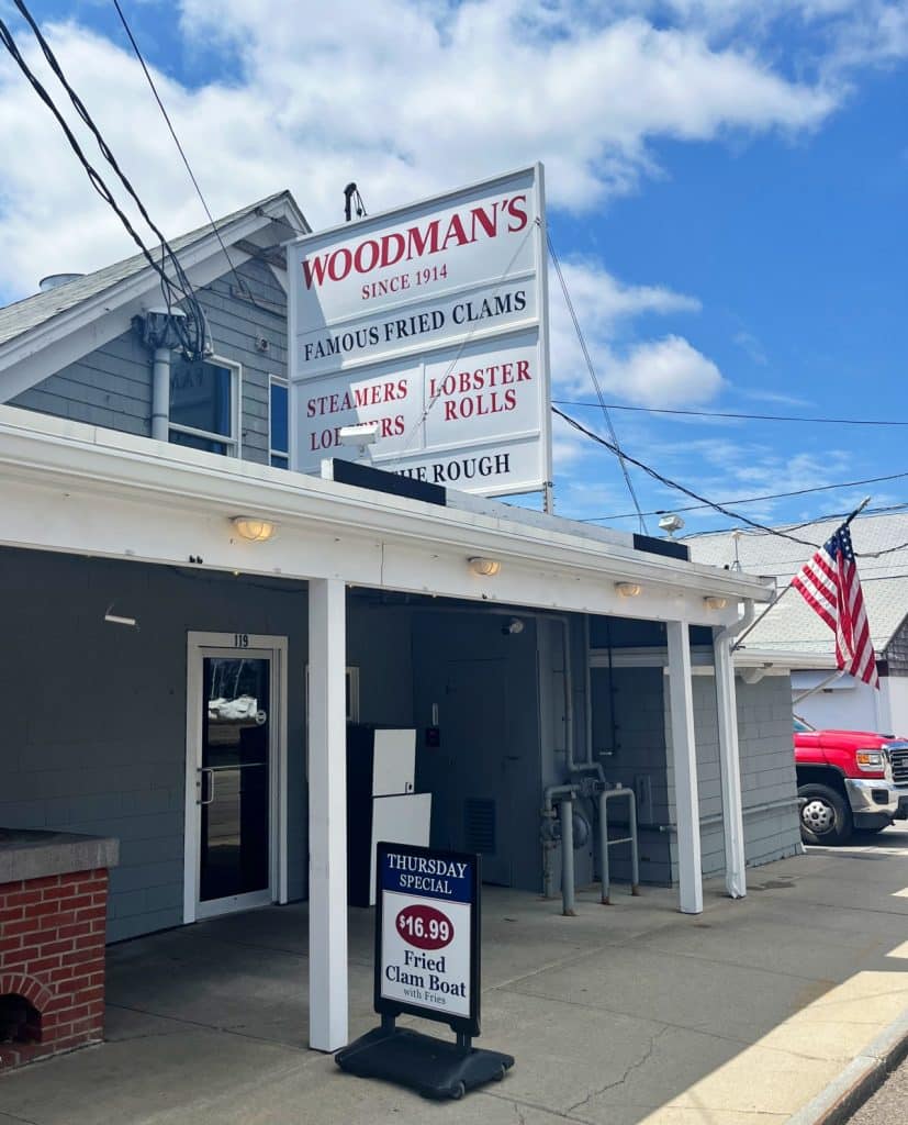 Front view of Woodman's of Essex restaurant with a sign advertising fried clams, steamers, and lobster rolls. A board on the sidewalk displays the Thursday special: Fried Clam Boat for $16.99.