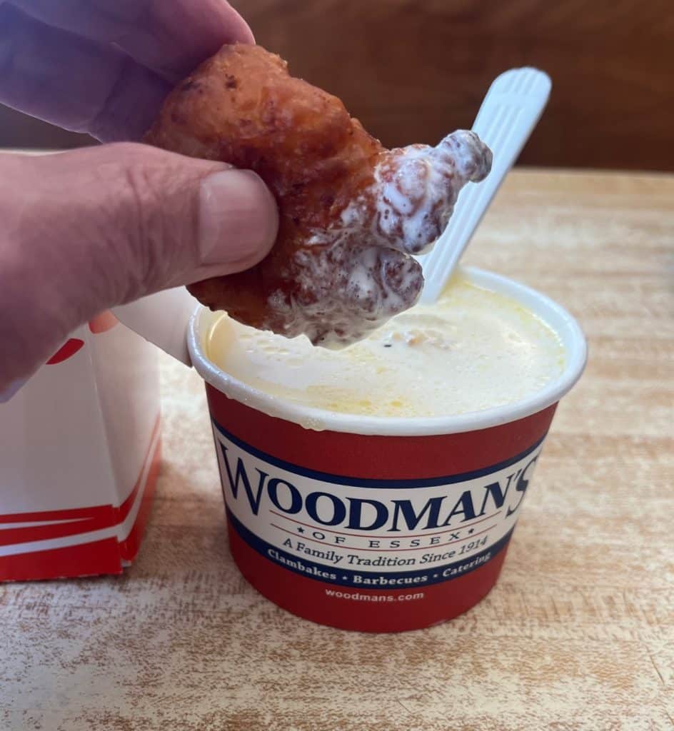 A hand dips a fried clam cake into a cup of creamy clam chowder from Woodman's of Essex.