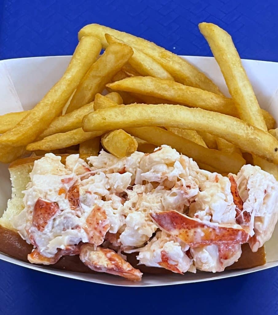 A serving tray from Woodman's of Essex, containing a lobster roll made with lobster meat in a mayo-based mix, accompanied by a side of golden French fries. The tray is placed on a blue surface.