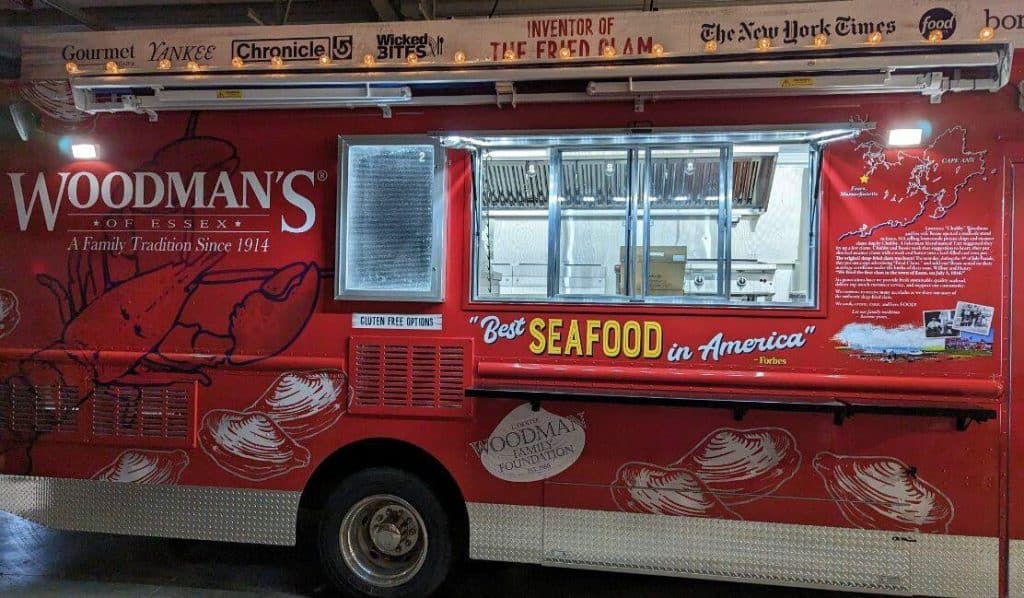 A red food truck with "Woodman's of Essex" logo, advertising "Best Seafood in America." The truck has various accolades and contact information painted on it.